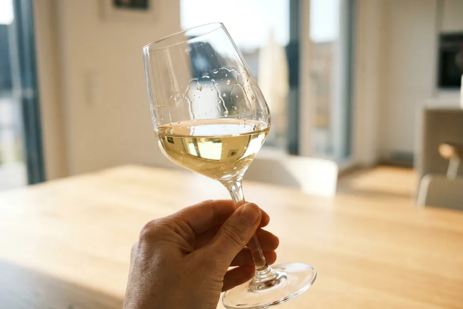 Hand hält ein geschwenktes Glas Weißwein gegen das Licht, Schlieren (Kirchenfenster) laufen an der Glasinnenwand herab